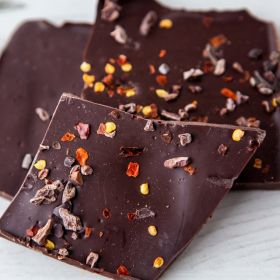 Dark Chocolate Oaky-smokey Chilli & Nib Slabs which are  vegan-friendly by Chococo on a wooden chopping board with chilli flakes in a white pot and pink linen napkin 